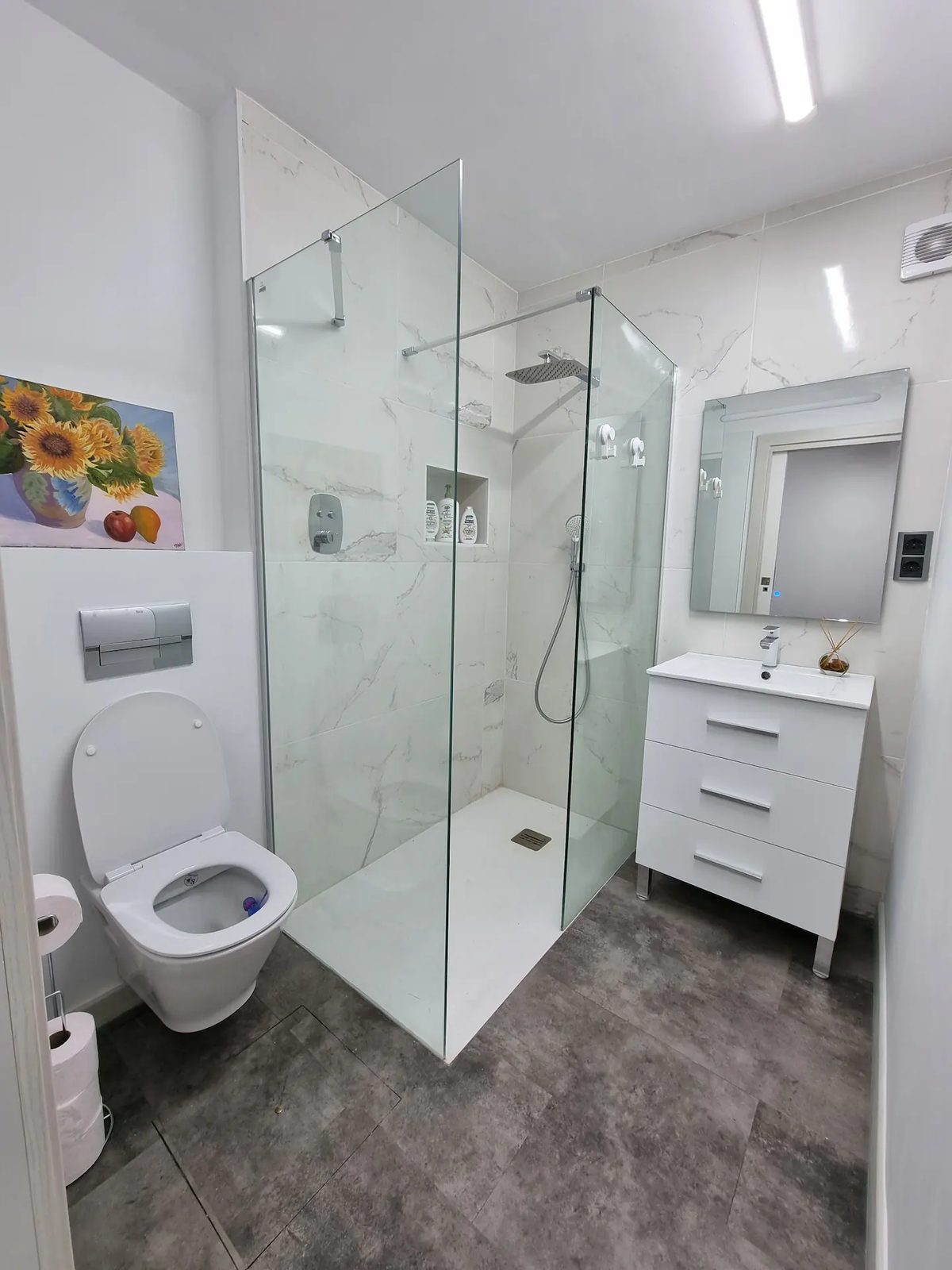 A walk in shower behind glass with marble tiling, beside a white vanity unit and mirror