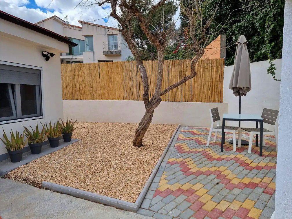 A walled garden with gravel, an olive tree, a patterned terrace and a table for two under a parasol