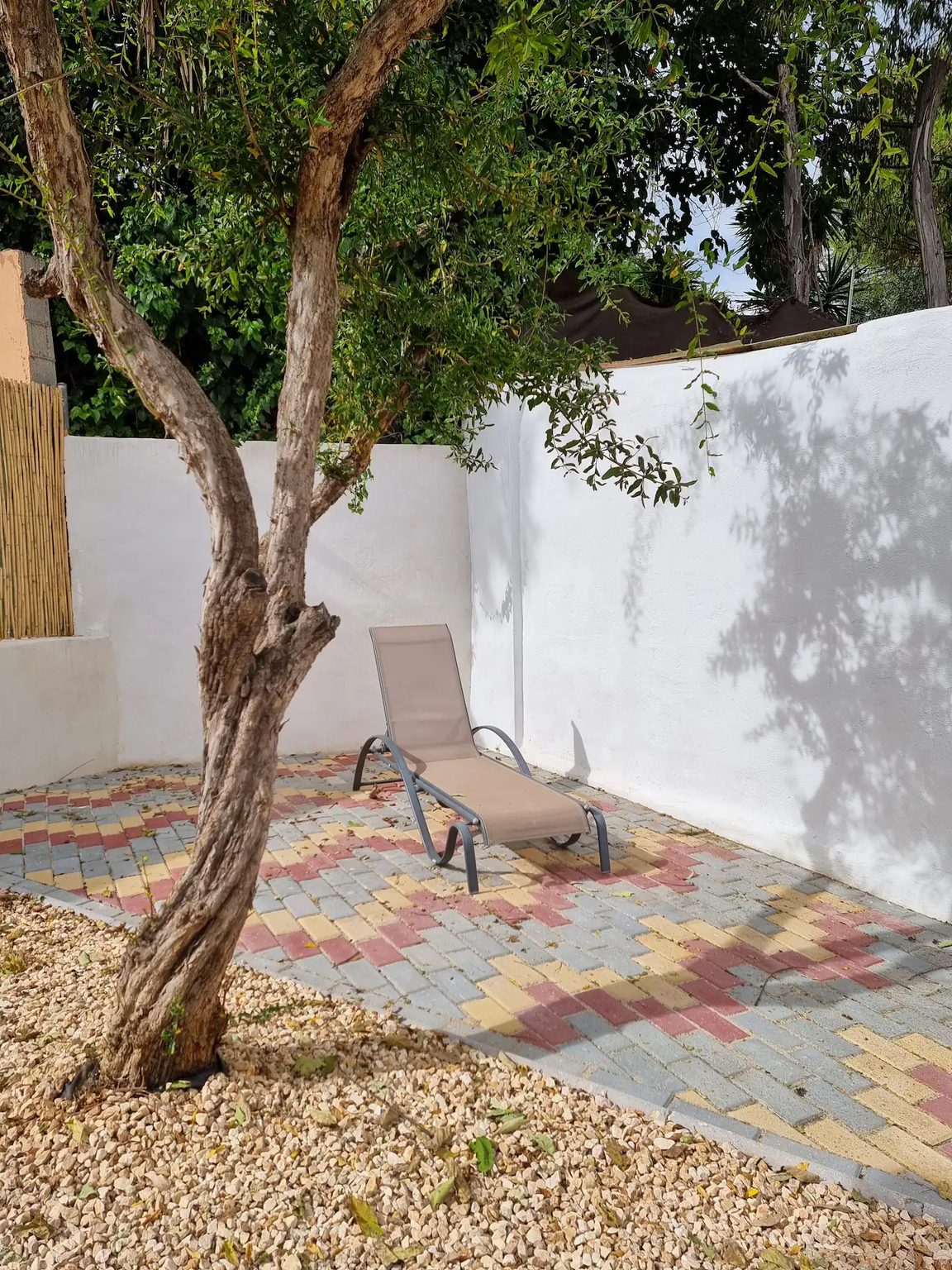 A sun lounger on a patterned terrace under a tree, against a white garden wall